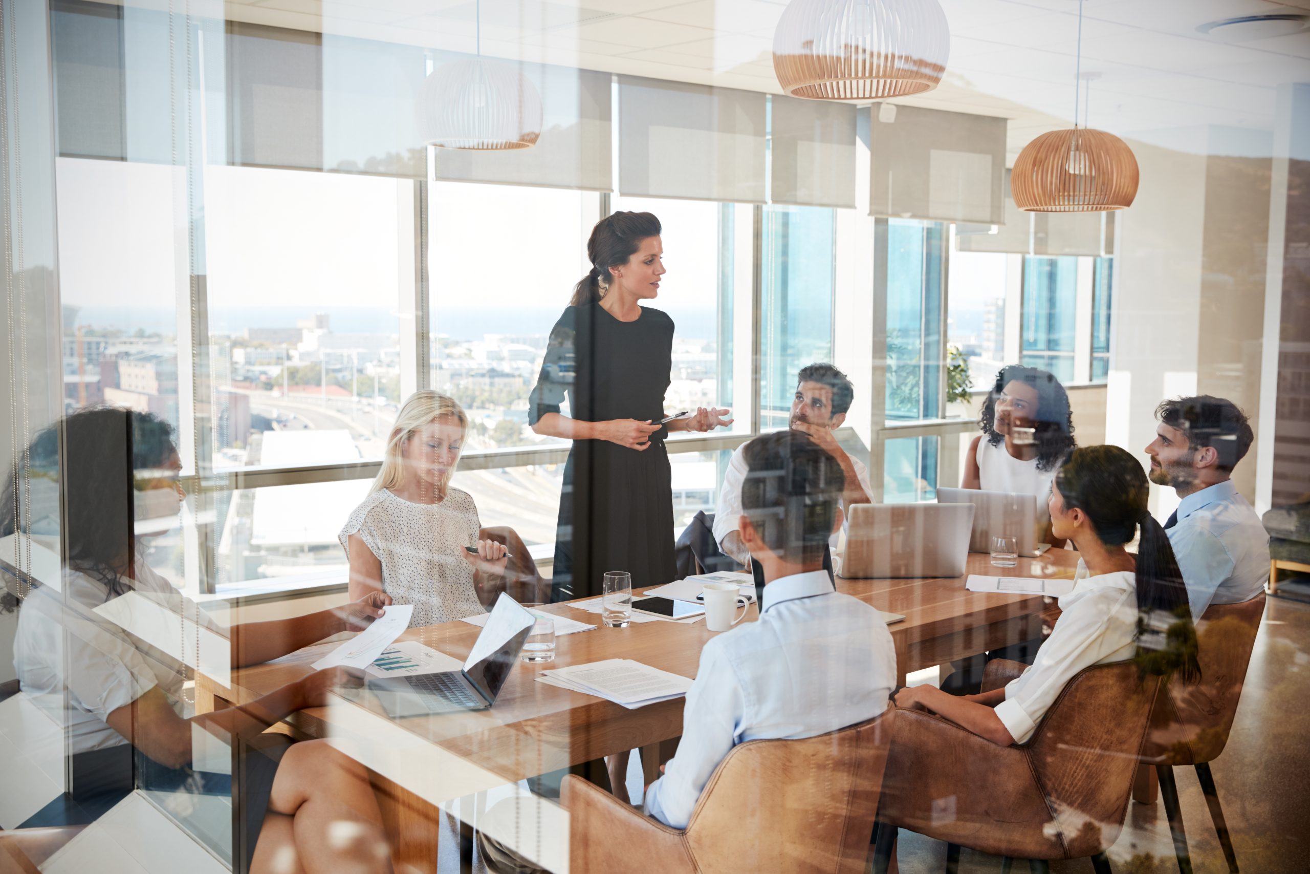 A leader stands to speak to a team around a table in a glass-walled meeting room